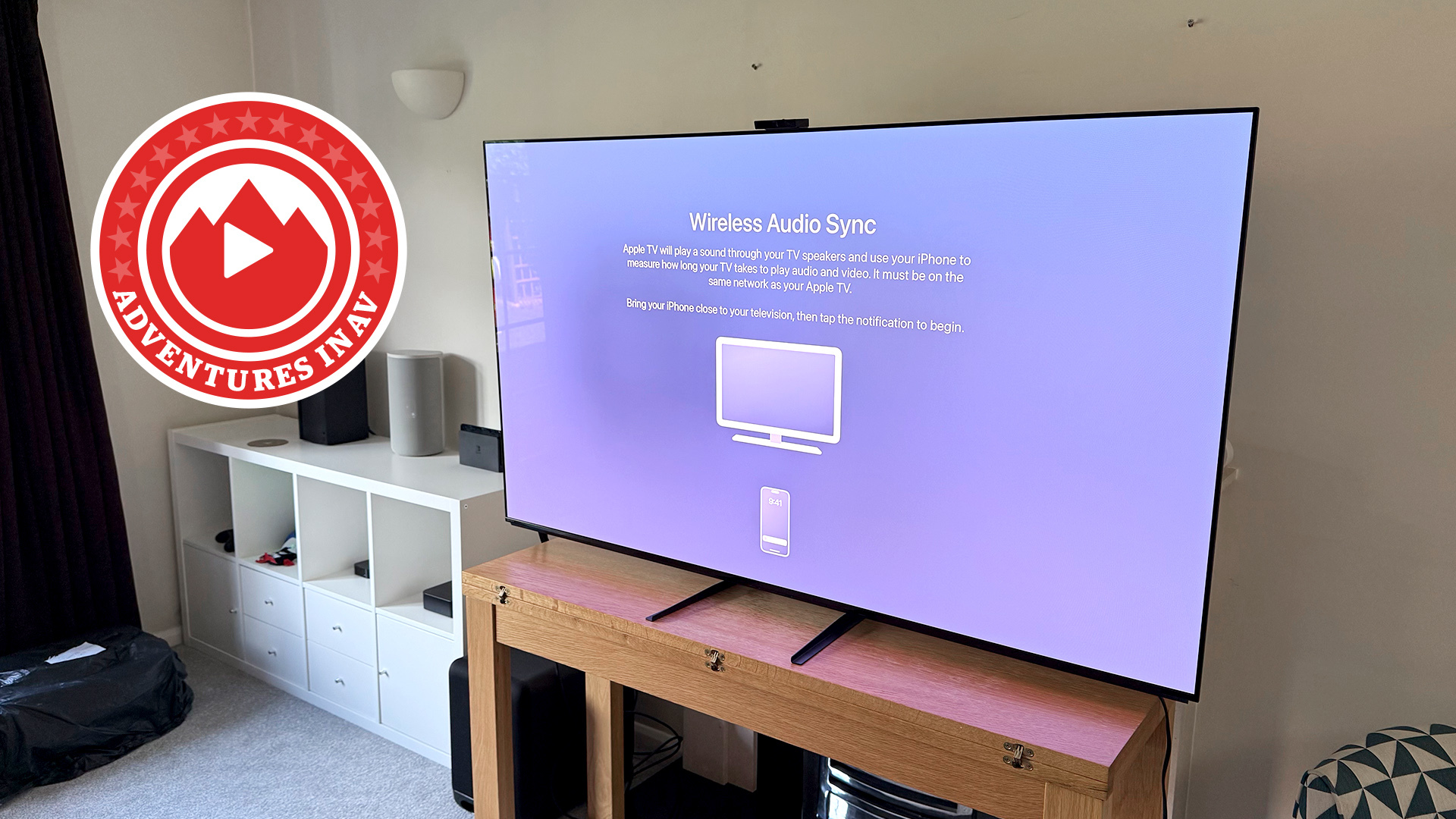 The Sony A95L, pictured on a wooden table in a living room. On the screen is the Wireless Audio Sync page of the Apple TV 4K. In the corner of the image is a red 'Adventures in AV' logo.