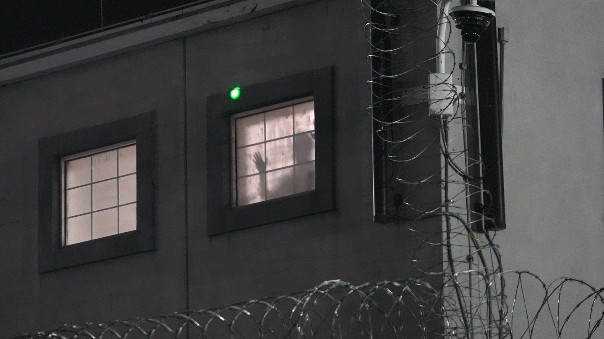 a detainee waves to protestors from a shadowy window. barbed wire surrounds the frame 