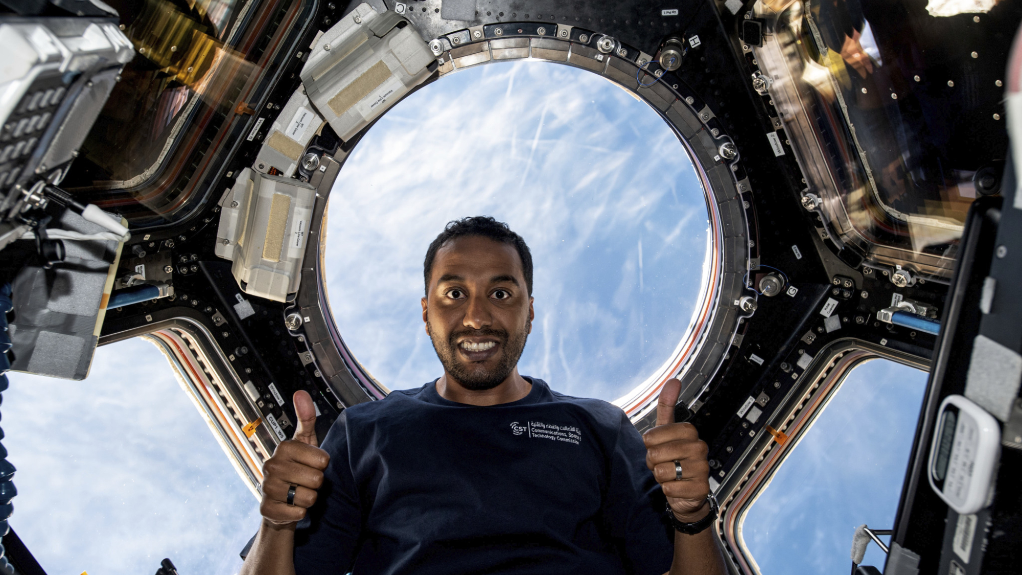 Saudi Space Commission astronaut Ali AlQarni with two thumbs up and the Earth through big windows behind him on the space station