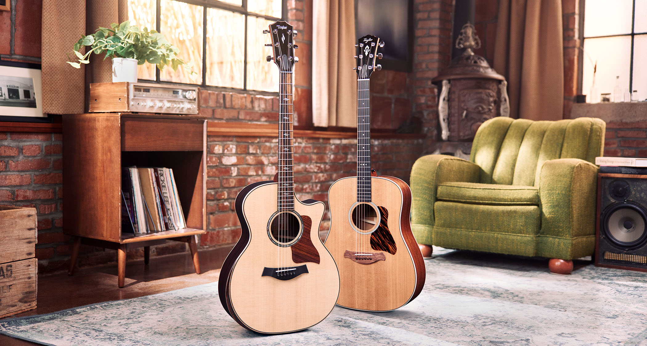 Taylor Guitars' new Next Generation Grand Auditorium cutaway acoustic is pictured with its new Gold Label Square-shouldered Dreadnought acoustic in a living room.