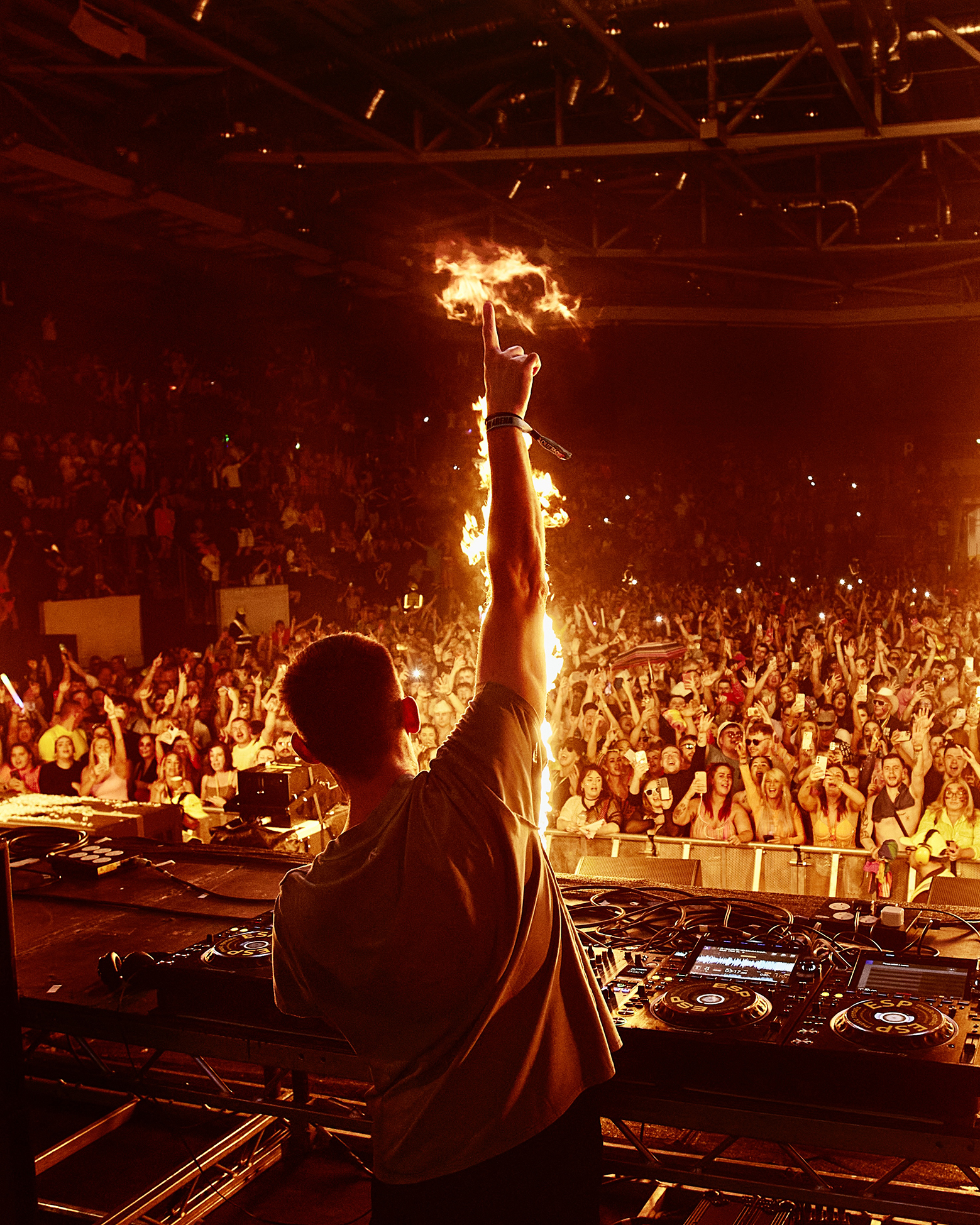 Darren Styles standing in front of the crowd at a live concert, raising his arm that is surrounded by fire effects from the gig