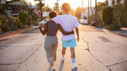 A young African American couple with their arms around each other walking down a suburban street at sunset shot from behind