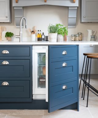 A bespoke Shaker-style kitchen with rich navy lower cabinets, fitted with brushed nickel cup handles, and light grey upper cabinets. The island features an integrated fridge drawer, complemented by a sleek stainless steel tap and a bright white worktop.