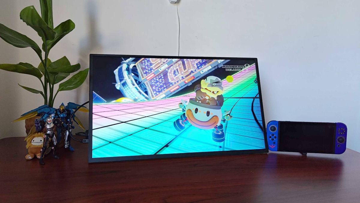 Uperfect Umax 24 portable monitor connected to Switch 2 next to plant, Pharah and Samus figures on woodgrain desk with Mario Kart 8 gameplay on screen.