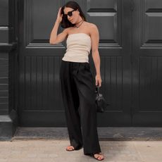 influencer Jessica Skye poses in front of a black wall in London wearing black sunglasses, a black cord necklace with a silver heart pendant, a beige ruched sleeveless top, black trousers, and a black bag