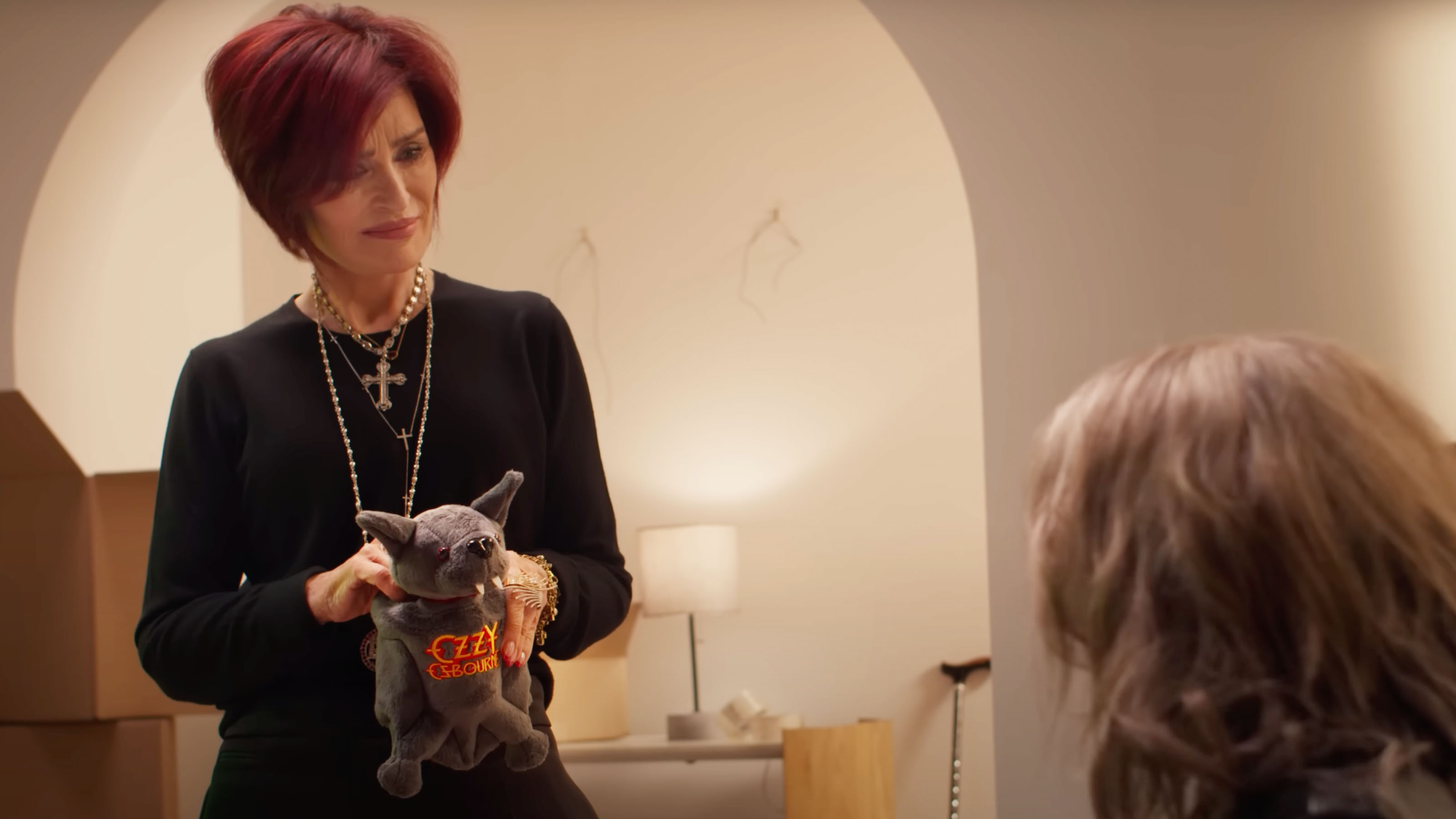 A shot of Sharon Osbourne in a brightly lit living area holding an Ozzy Osbourne branded bat plush