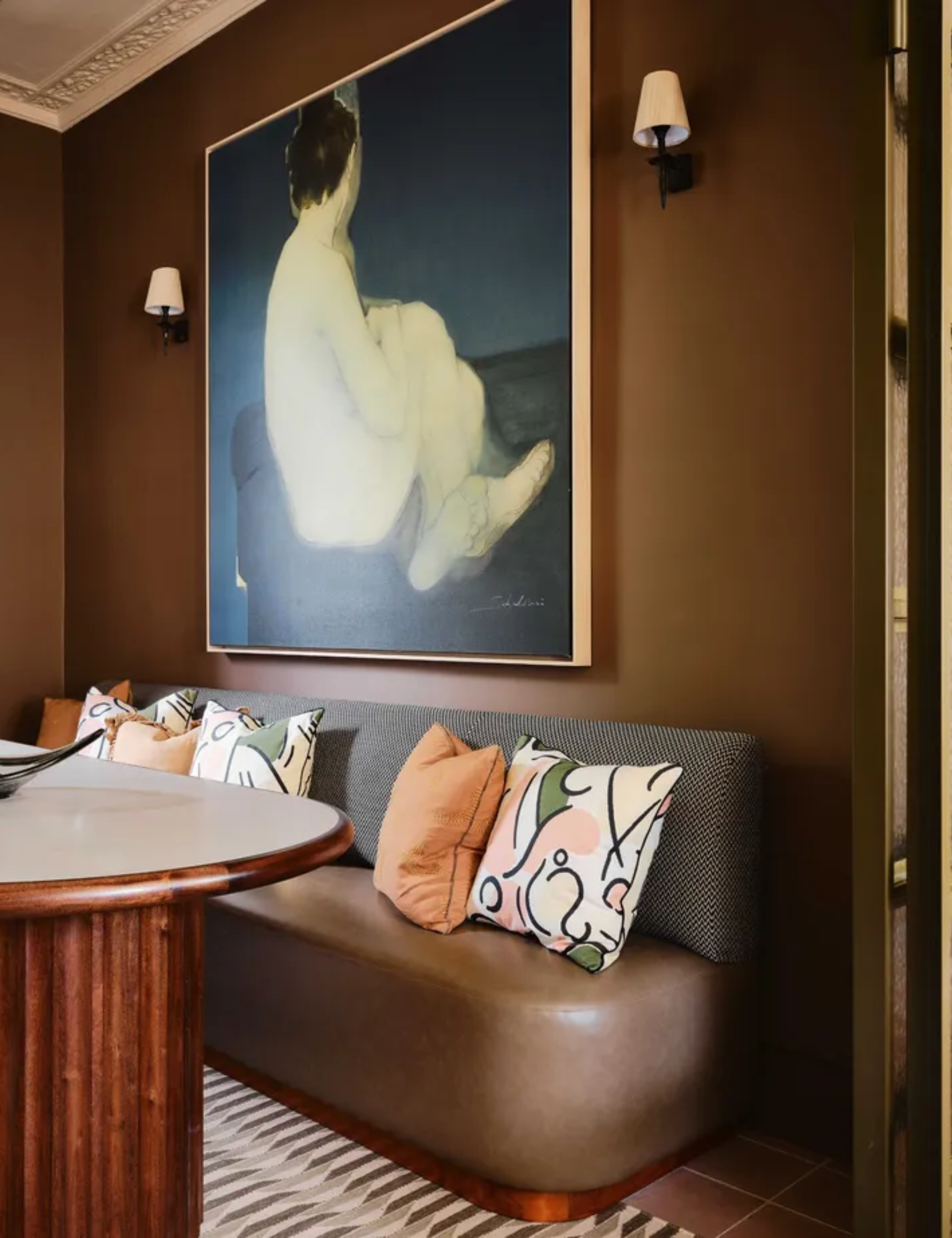 A brown dining room with a curved wooden table with a fluted base, and cozy upholstered banquette, and a large framed painting on the wall