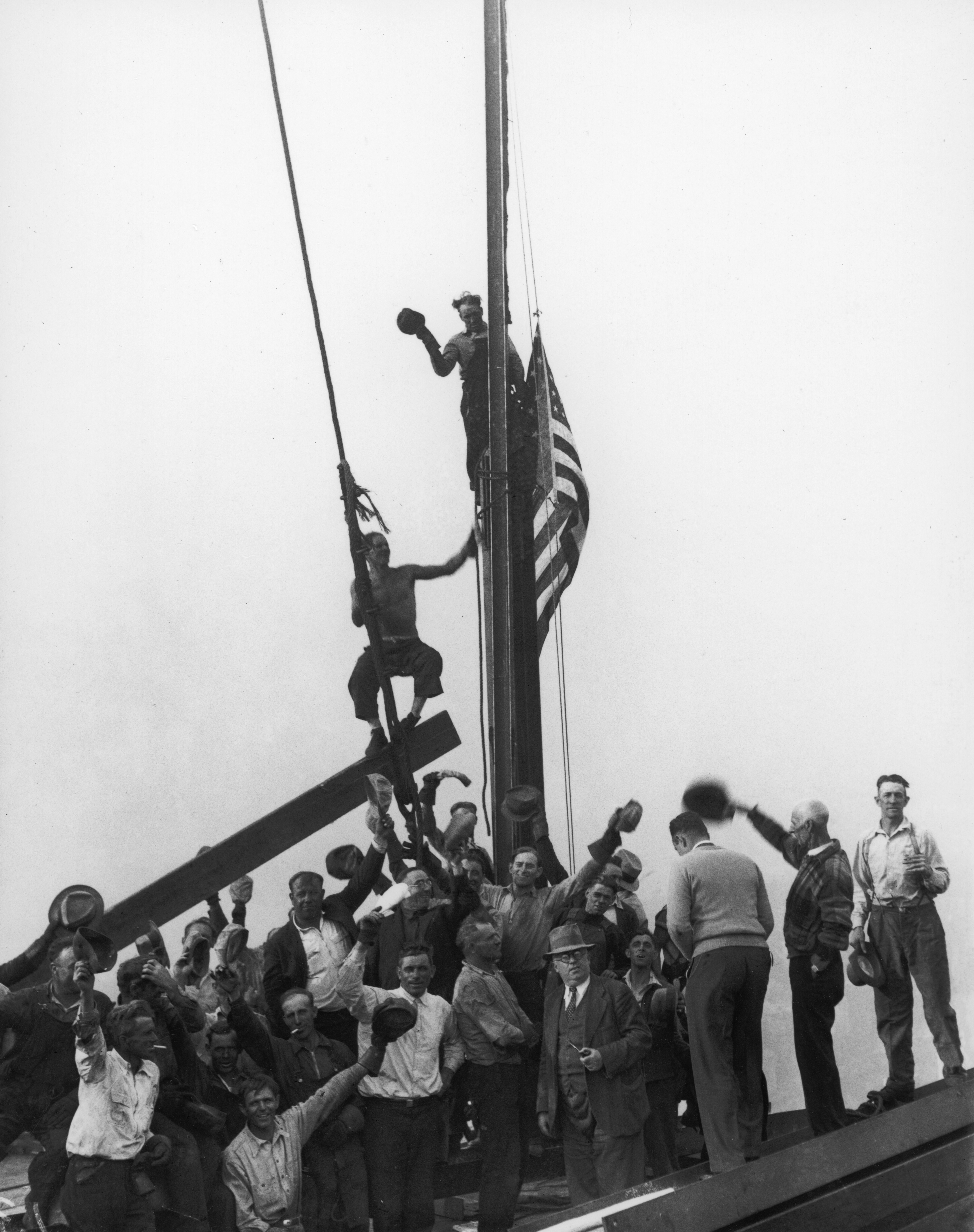 Workers crowd a high steel platform, waving their hats and cheering as two ironworkers cling to a flagpole from which an American flag has just been raised to mark the building's structural completion.