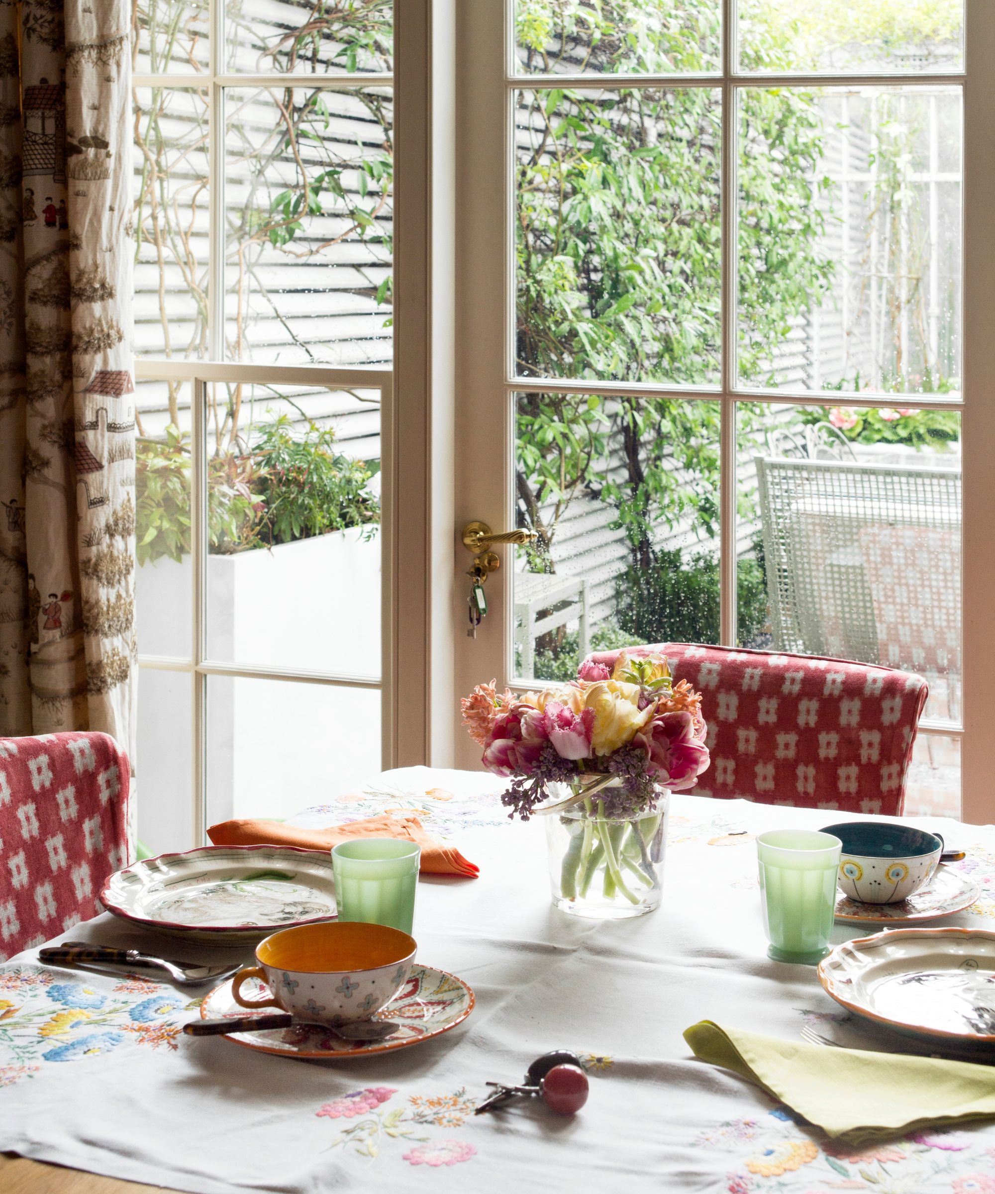 Dining room with French doors looking out onto the garden. The dining table is decorated with red patterned chairs, tablecloth, colourful napkins, and china