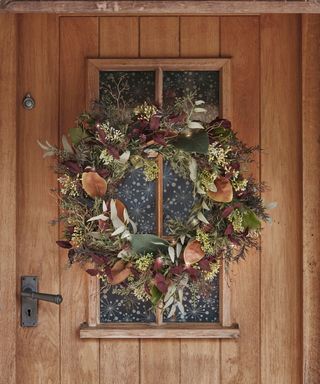 A wooden front door with an autumn wreath made of sage green and orange leaves