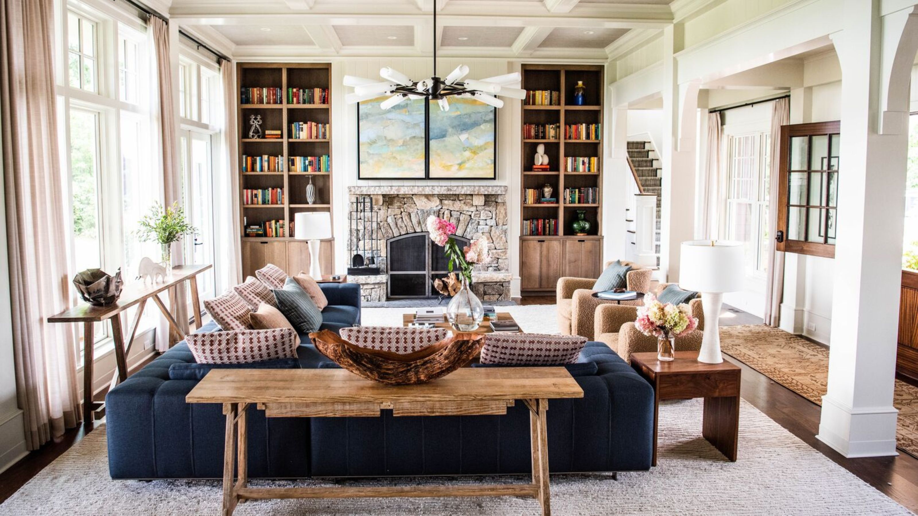 large neutral living room with large windows, a stone fireplace with wooden built-ins in the alcoves, a large navy velvet couch with a rustic wooden sofa table and a huge rug