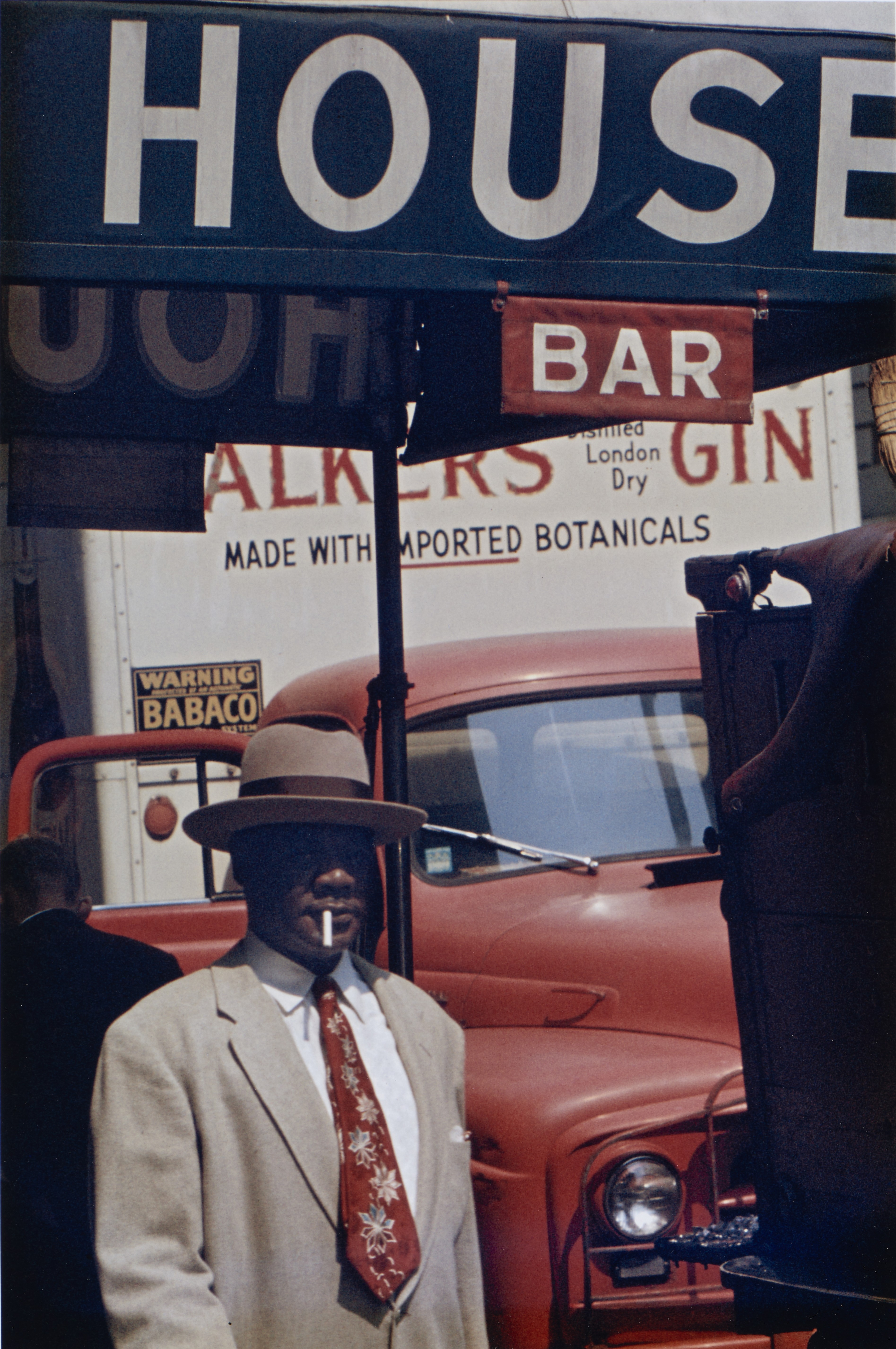A man in a cream suit, patterned tie and wide-brimmed hat stands beneath a "House Bar" sign with a cigarette at his lip, a red delivery truck filling the frame behind him.