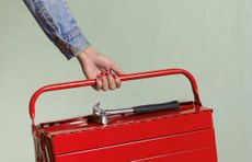 Woman with red fingernails and denim shirt holding red toolbox with hammer sitting on top