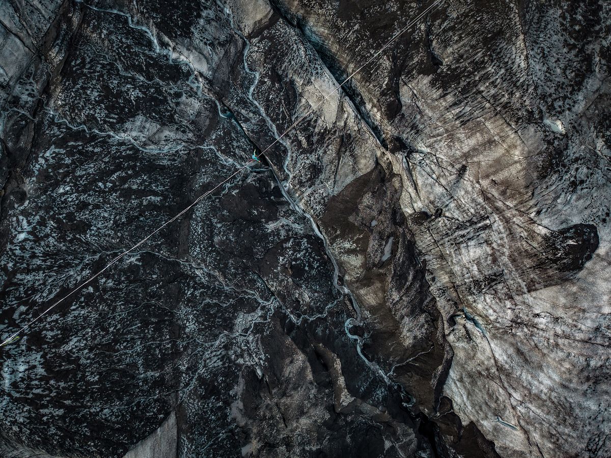 Aerial view of a climber suspended on a rope over dark, rugged glacier ice with deep crevasses