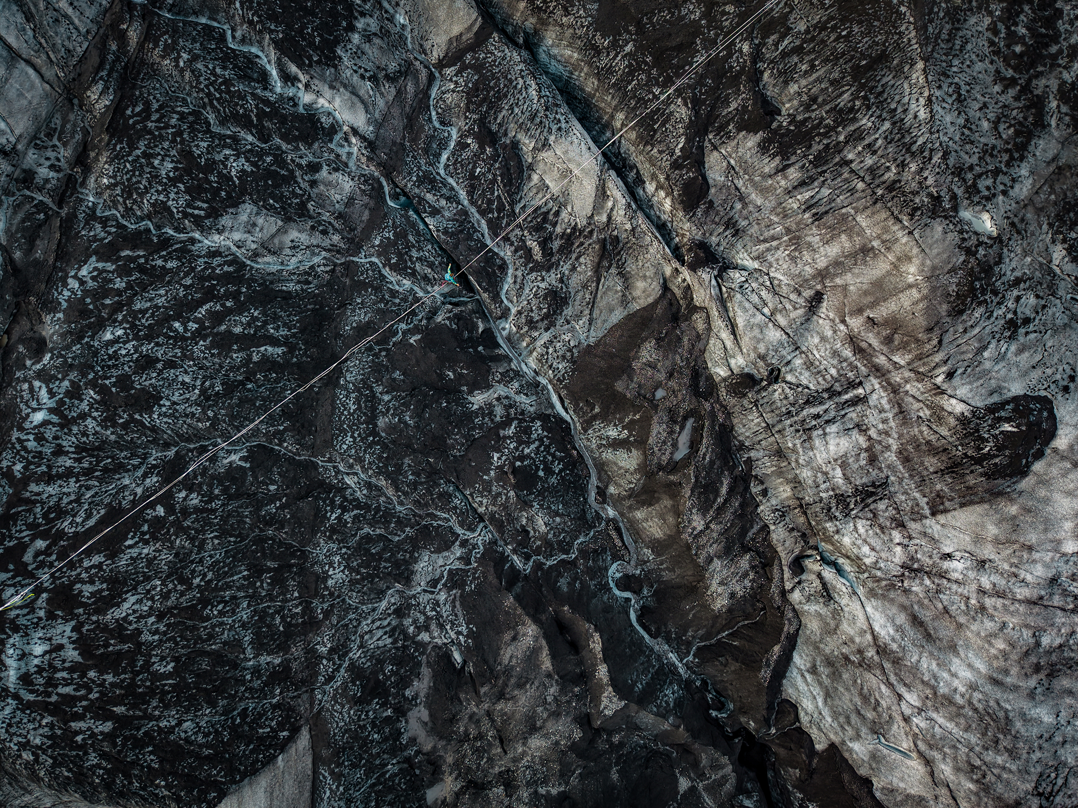 Aerial view of a climber suspended on a rope over dark, rugged glacier ice with deep crevasses