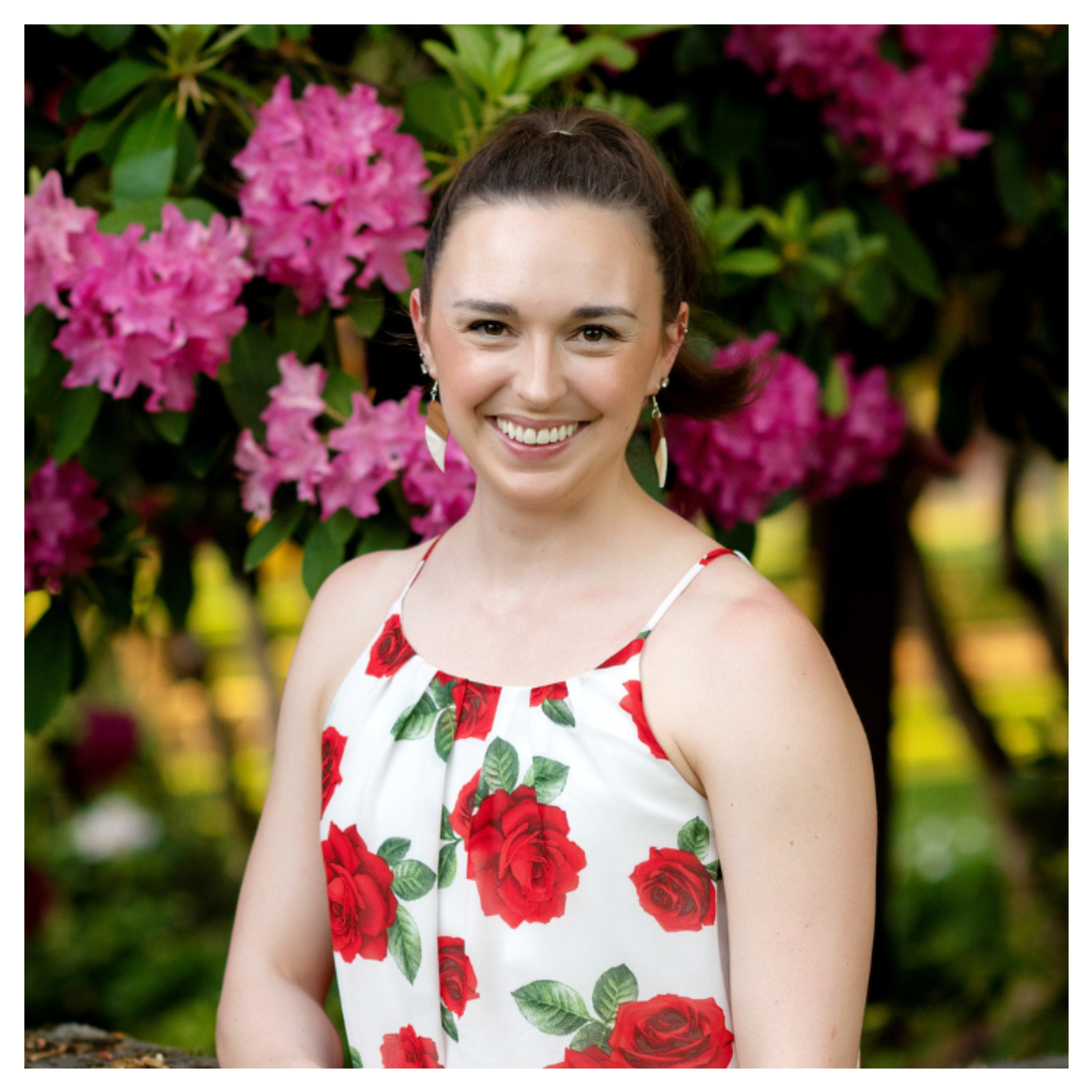 A woman smiling in front of a tree with pink flowers in a white spaghetti strap top with red roses and green leaves