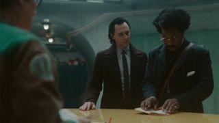 Image from the Marvel T.V. show Loki, season 2 episode 4. In front of a wooden desk are two men wearing suits. The man on the left has dark slicked back hair. The man on the right has thick dark hair, round, gold glasses and is holding opening an orange notebook on the desk. Behind the desk we can see the side of another man who is wearing black glasses and a jumpsuit.