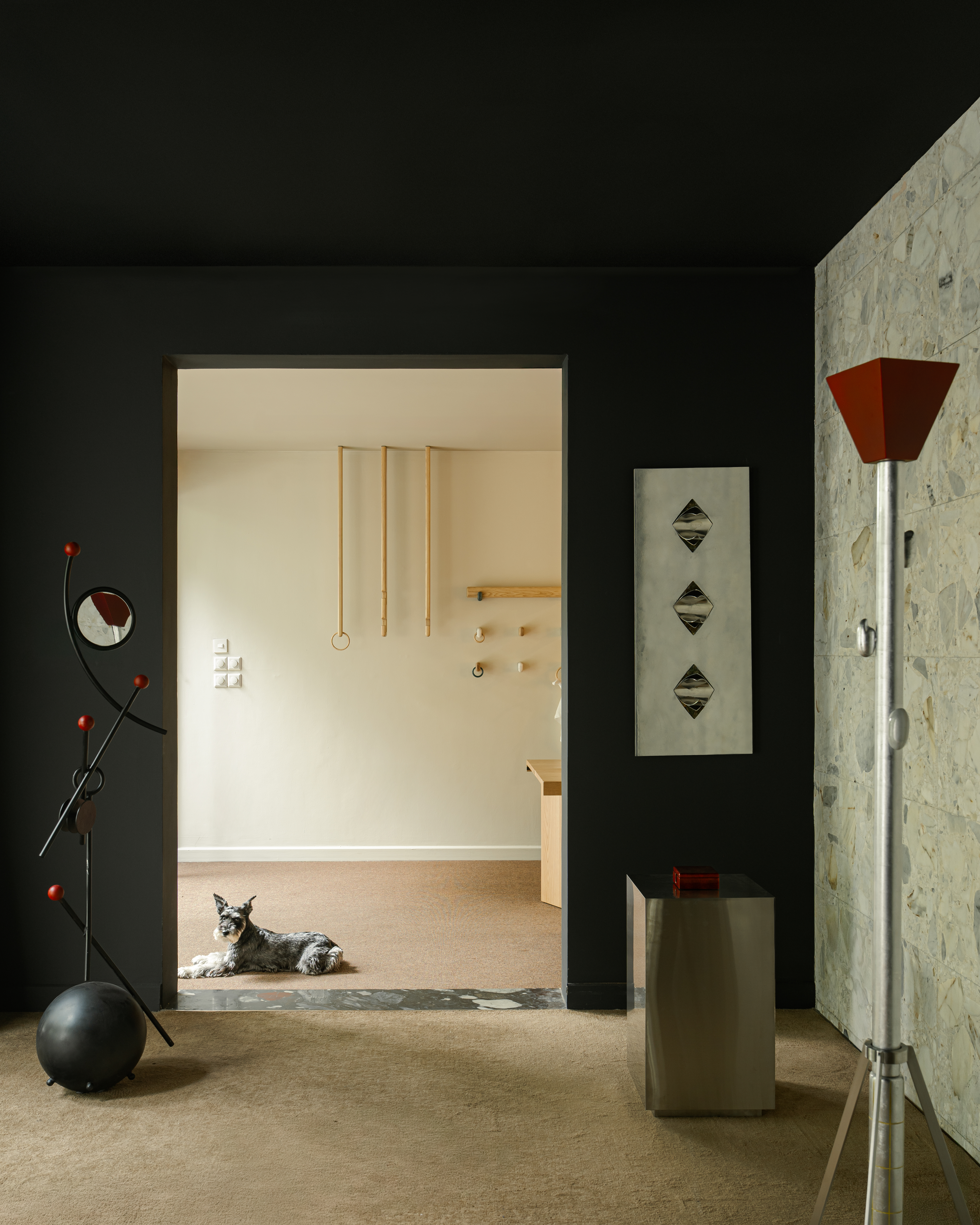 A beautifully decorated gallery with half black, half painted, half rock walls is filled with geometric sculptures in wrought iron, wood, and chrome, as a dog sits on its neutral floor.
