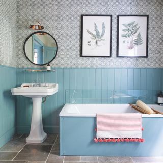 bathroom with half panelled blue walls and wallpaper above with bath and basin and pictures on the wall