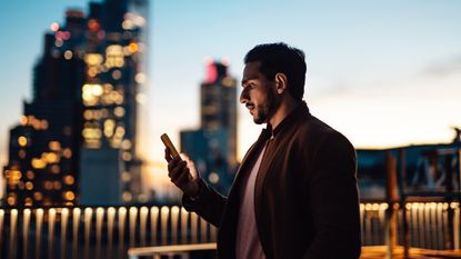 A young businessman looks at his phone while standing on the roof of a skyscraper with a big-city skyline in the background at dusk.