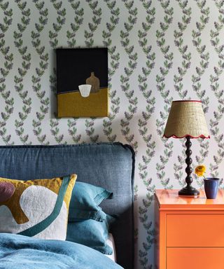 Pretty small scale bedroom ideas showing botanical print wallpaper on bedroom wall with blue upholstered linen headboard and vibrant orange bedside cabinet