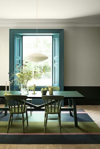 Open full length blue shutters framing white lantern ceiling pendant, color block striped rug, and fresh meadow arrangement on table.