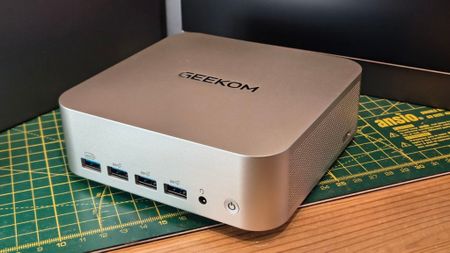 A sleek, compact Geekom mini PC with a silver finish sits on a green cutting mat. It features USB ports and a power button, conveying a modern tech vibe.