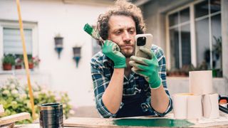 male in the middle of painting wood outside but pausing for a break and looking at his phone