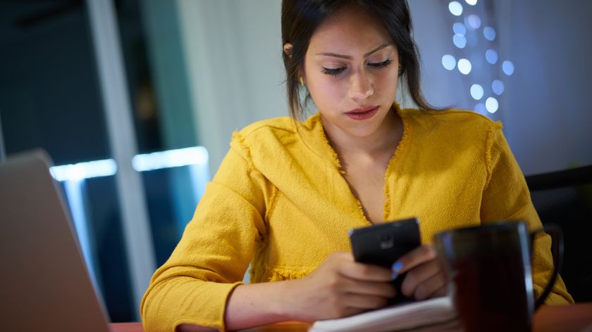 A woman sitting at a table while using a phone with. She also has a mug near her. 