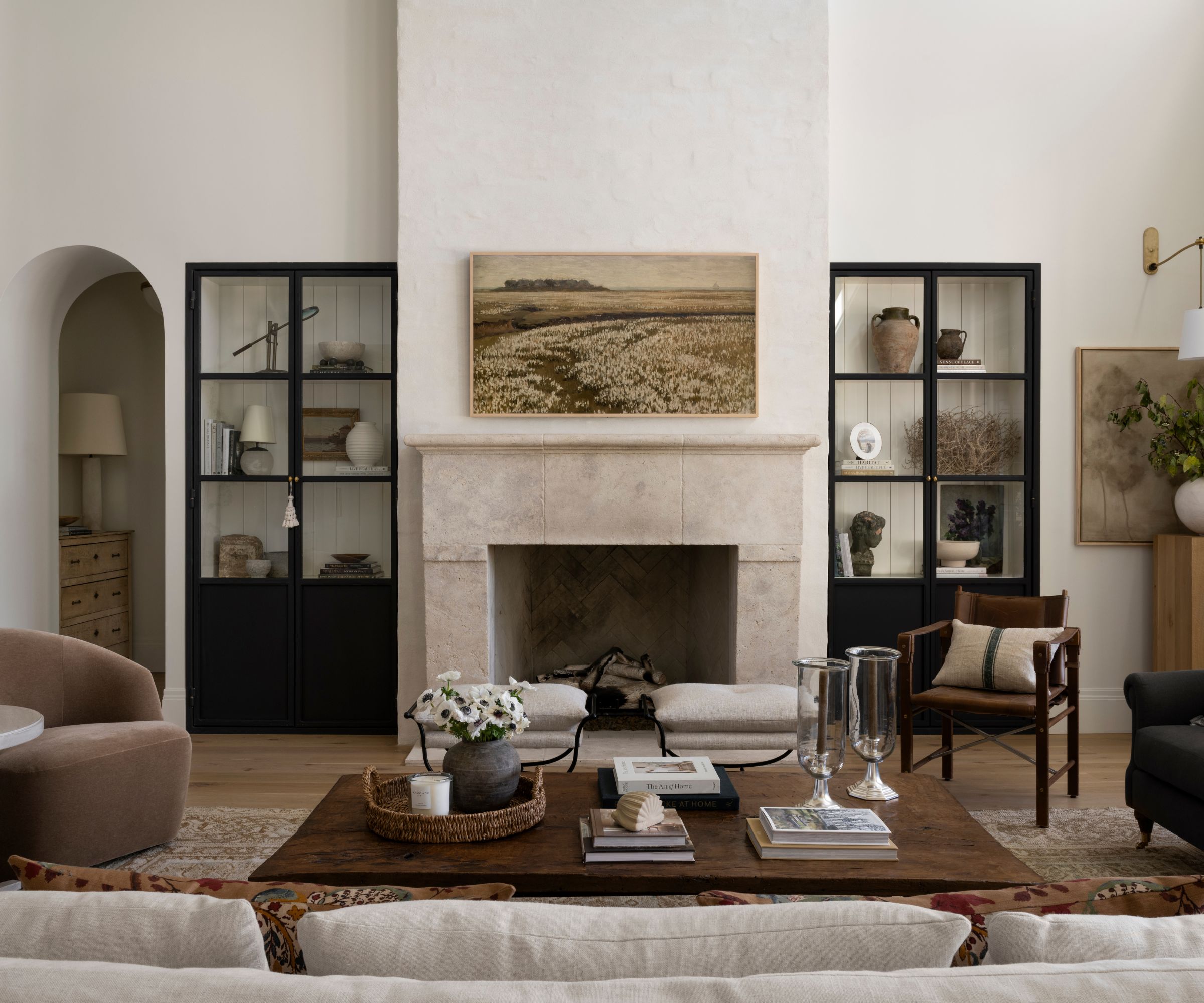 Transitional living room with high ceilings, walls painted in Benjamin Moore's Swiss Coffee, a stone fireplace, a coffee table holding books and trinkets, and glass-fronted cabinets filled with decor.