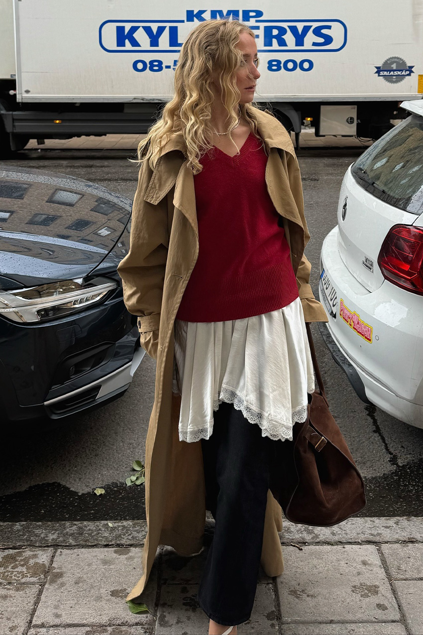 A sweater styling trend is shown in a photo of a woman&#039;s outfit where she&#039;s standing on the sidewalk in front of parked cars wearing a long tan trench coat on top of red v-neck sweater that&#039;s layered over a white slip dress and black jeans styled with white cutout heels and a chocolate brown suede bowler bag