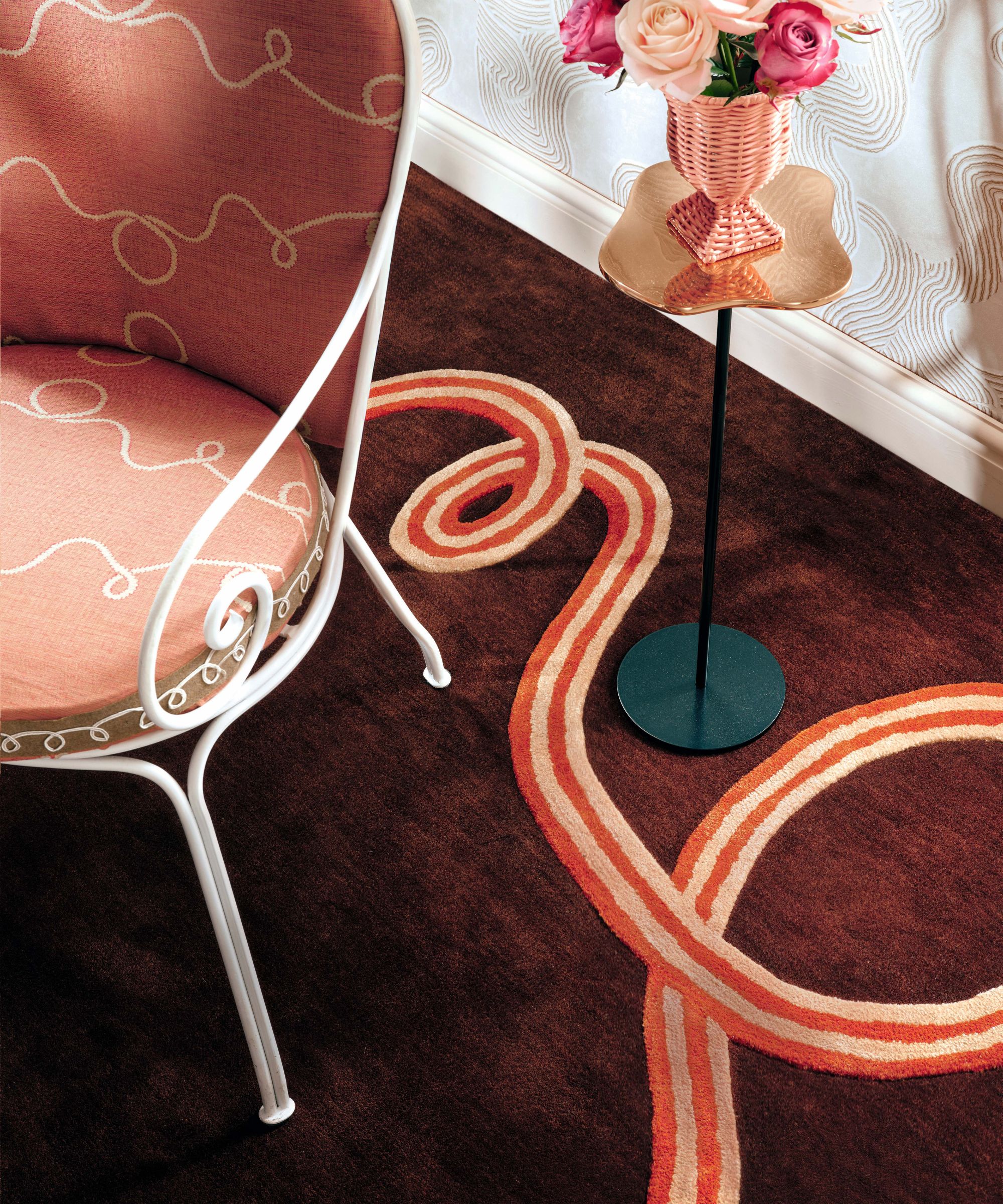 A high-angle shot of a whimsical interior featuring a chocolate-brown rug with a bold, winding orange-and-cream ribbon pattern. A white wrought-iron chair with salmon-pink patterned cushions sits to the left, and a slender gold pedestal table holding a pink wicker vase of roses stands against a wavy-patterned wallpaper.