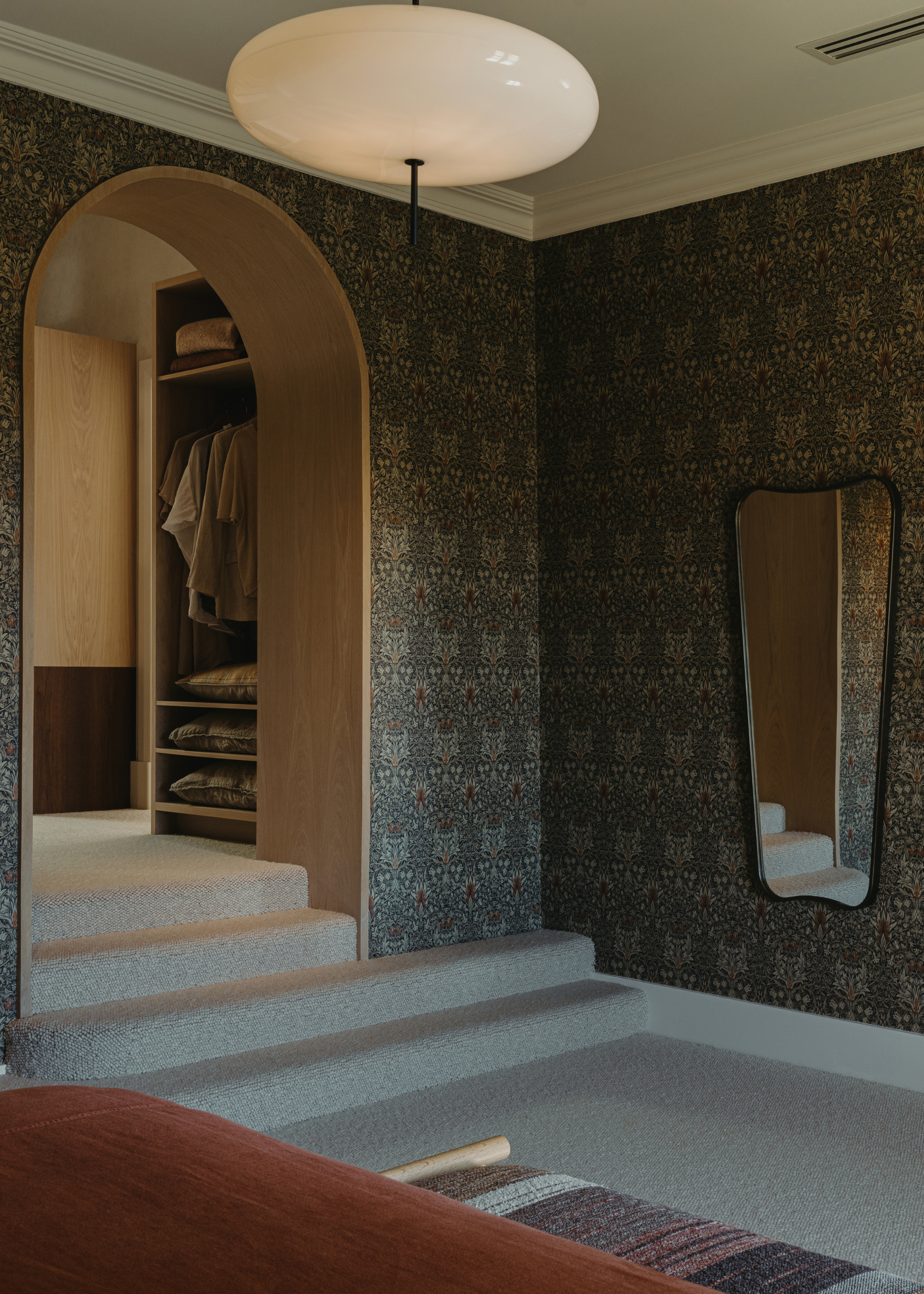 A bedroom with patterned wallpaper, a hanging lamp, a mirror, an arched doorway leading to a wardrobe