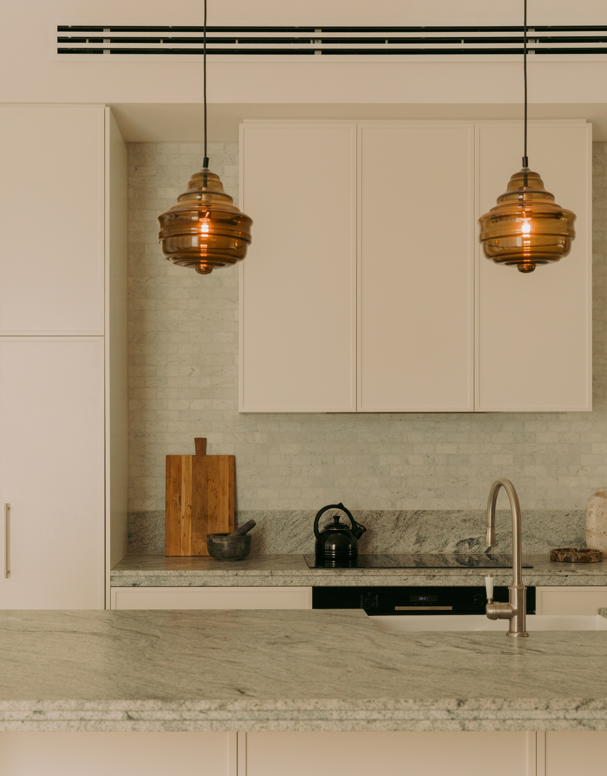 A minimalist contemporary kitchen with a green marble island, a green tiled backsplash, white cabinets, and amber pendants