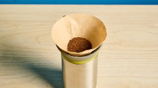 Wacaco Cuppamoka, a stainless steel coffee pour-over brewer, photographed against a blue background, with brown filters and a sippy lid