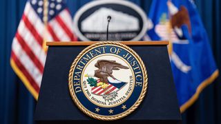 LockBit: A podium bearing the seal of the Department of Justice (an eagle carrying the US flag and a sprig of leaves) in the foreground, with two flags out of focus in the background (the US on the left, a flag bearing the seal on the right).