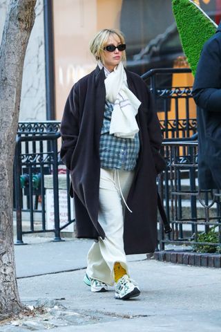 Jennifer Lawrence walking on the sidewalk in New York City wearing a black coat by The Row, a tied scarf, a plaid blue button-down shirt, lounge pants, yellow Polo socks, and New Balance 1906 sneakers.