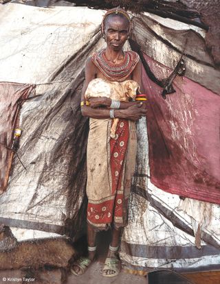 An elderly Rendille woman holds her newborn goat outside her houes