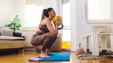 woman in a deep squat holding a yellow kettlebell at her chest, on a blue exercise mat in a living room setting.