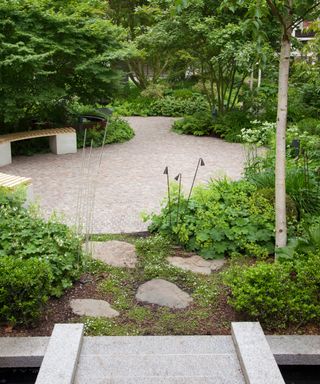 A round brick patio design in a wooded garden with stepping stones and a stone bridge.
