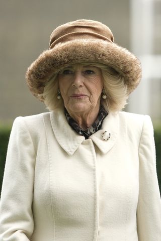 Queen Camilla wearing a white coat and tan fur hat with a serious expression