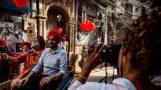 A female photographer placed to the right is photographing three men with red and black turbans sitting on chairs with red balloons in the background