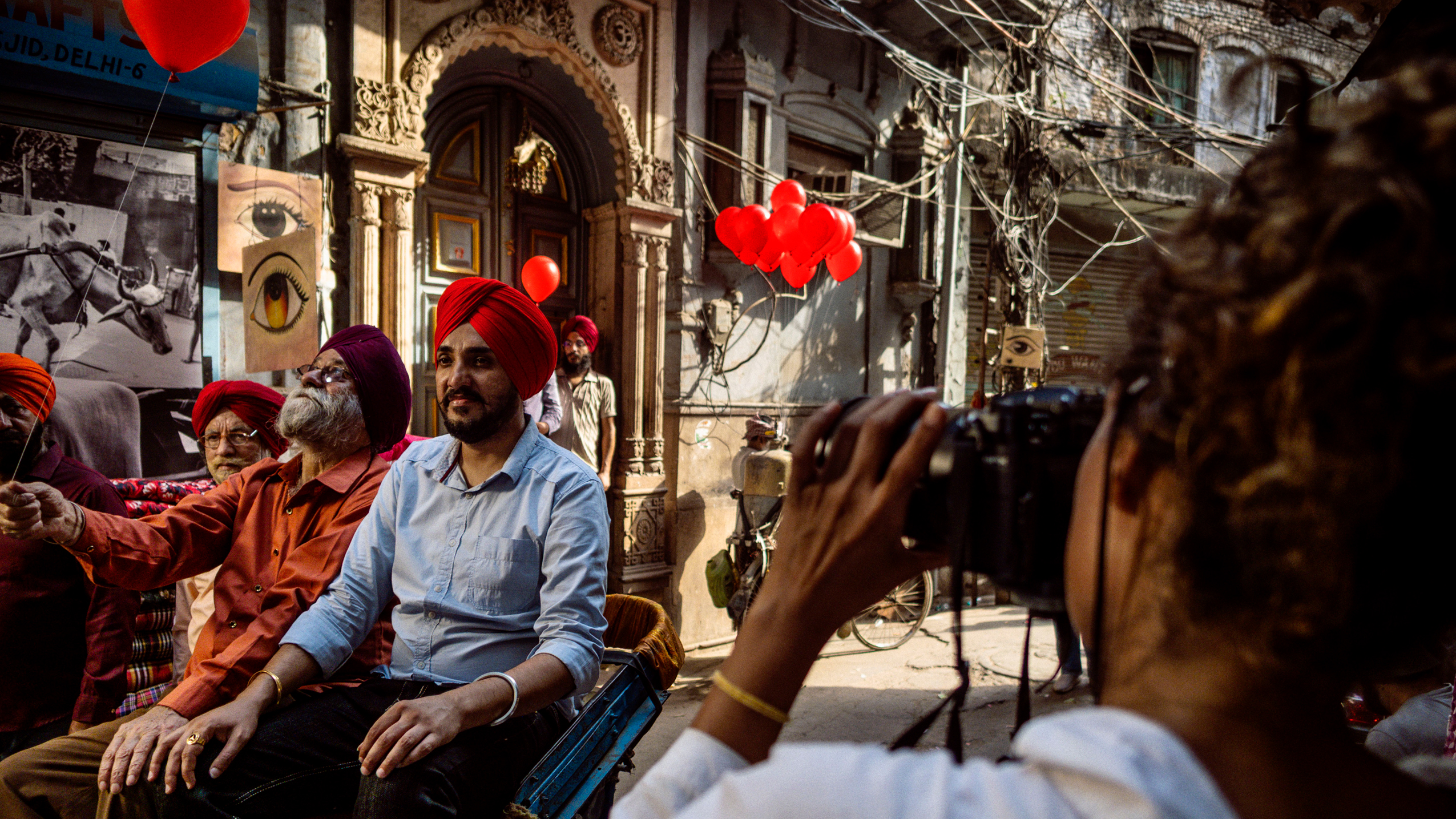 A female photographer placed to the right is photographing three men with red and black turbans sitting on chairs with red balloons in the background