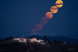 super blue moon appears to rise up behind wind turbines with a small village in the foreground.