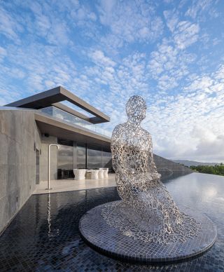 Large grey metal sculpture of a man kneeling down surrounded by infinity pool
