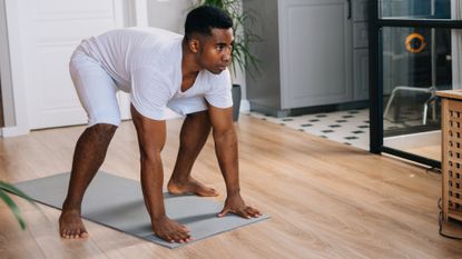 man mid way through a burpee in a half squat position wearing white tshirt and white shorts in his flat