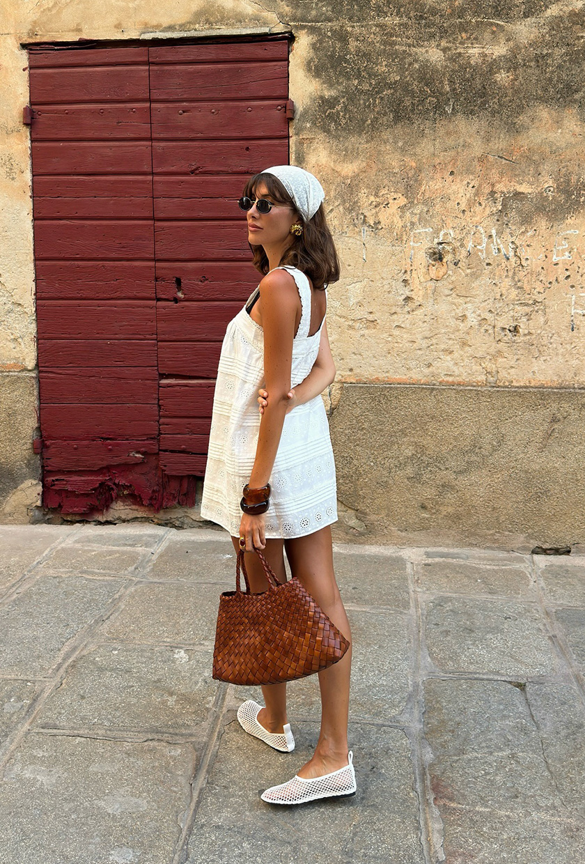 The summer aesthetic, coastal boho is shown in a street style photo of a woman standing on the sidewalk wearing a white headscarf with black sunglasses, gold earrings, a white eyelet mini shift dress, stacked brown bangles, a brown woven leather tote bag, and white mesh ballet flats