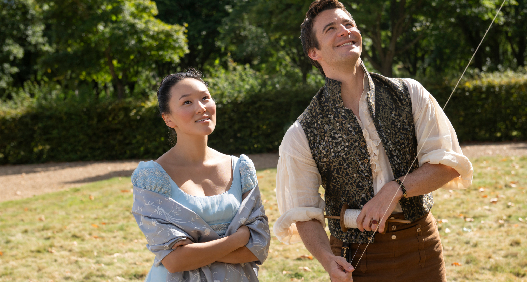yerin ha as sophie baek wearing a blue dress and luke thompson as benedict bridgerton flying a kite in a sunny field in bridgerton season 4