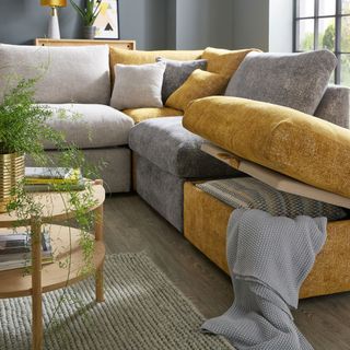 a living room with light grey, dark grey and orange couches with lift up storage, a brown circle coffee table with pot plant and books on top