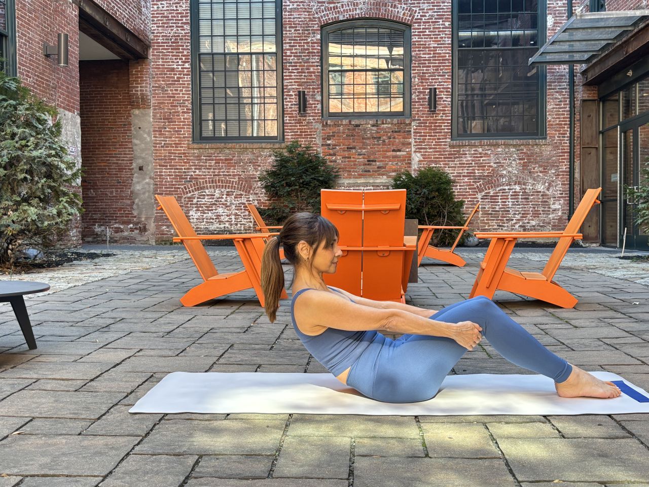 Pilates instructor Monica Borodin performs a half roll down. She wears a vest and leggings and is exercising outside on a mat. Behind her we see a brick building with large windows and table and chairs. She is seated, with her knees bent and feet on the floor. Her hands clasp the back of her thighs and she is leaning backwards.